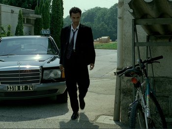 Movie still from “Oh, Woe Is Me” (1993), directed by Jean-Luc Godard – A man in a suit and tie walking down a street; Wide shot, Low angle