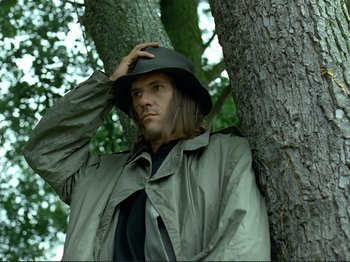 Movie still from “Oh, Woe Is Me” (1993), directed by Jean-Luc Godard – A man wearing a rain coat and hat leaning against a tree; Close Up shot, Low angle