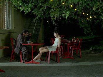 Movie still from “Oh, Woe Is Me” (1993), directed by Jean-Luc Godard – A man and a woman sitting at a table under a tree; Wide shot, High angle