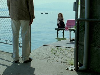 Movie still from “Oh, Woe Is Me” (1993), directed by Jean-Luc Godard – A woman sitting on a bench on a pier; Wide shot, Over the shoulder angle