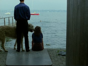 Movie still from “Oh, Woe Is Me” (1993), directed by Jean-Luc Godard – A man and a woman sitting on a dock next to a body of water; Wide shot, Over the shoulder angle