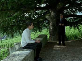 Movie still from “Oh, Woe Is Me” (1993), directed by Jean-Luc Godard – A man sitting on top of a bench next to a tree; Wide shot, Over the shoulder angle