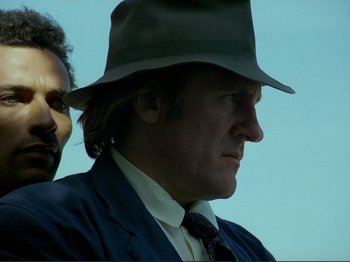 Movie still from “Oh, Woe Is Me” (1993), directed by Jean-Luc Godard – A person wearing a suit and a hat; Close Up shot, Low angle