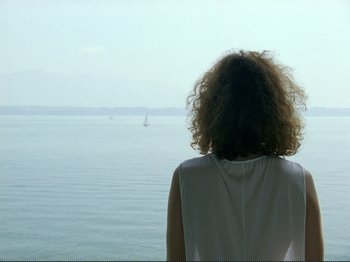 Movie still from “Oh, Woe Is Me” (1993), directed by Jean-Luc Godard – A woman with curly hair looking out at the ocean; Medium shot, Over the shoulder angle