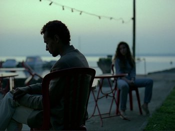 Movie still from “Oh, Woe Is Me” (1993), directed by Jean-Luc Godard – A man and a woman sitting at a table outside; Medium shot, Over the shoulder angle