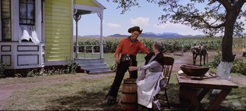 Movie still from “Oklahoma!” (1955), directed by Fred Zinnemann – A man and a woman sitting on a barrel in a yard; Wide shot, Over the shoulder angle