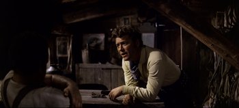 Movie still from “Oklahoma!” (1955), directed by Fred Zinnemann – A man leaning on a wooden table in a room; Medium shot, Over the shoulder angle
