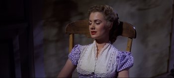 Movie still from “Oklahoma!” (1955), directed by Fred Zinnemann – A woman sitting in a wooden chair wearing a purple and white dress; Medium shot, High angle