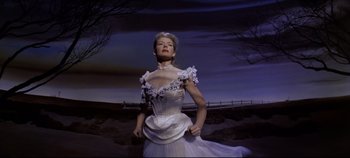 Movie still from “Oklahoma!” (1955), directed by Fred Zinnemann – A woman in a white dress standing in front of a fence; Medium shot, Low angle