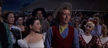 Movie still from “Oklahoma!” (1955), directed by Fred Zinnemann – A man and a woman are standing in front of a crowd; Medium shot, Low angle