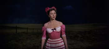 Movie still from “Oklahoma!” (1955), directed by Fred Zinnemann – A woman in a pink and white dress standing in a field; Medium shot, Low angle