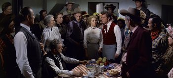 Movie still from “Oklahoma!” (1955), directed by Fred Zinnemann – A group of people standing around a table; Medium shot, Over the shoulder angle