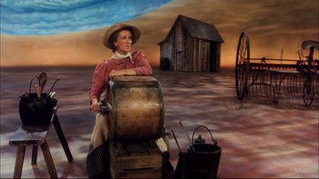 Movie still from “Oklahoma!” (1999), directed by Trevor Nunn – A woman sitting on top of a barrel in a field; Wide shot, High angle