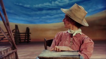 Movie still from “Oklahoma!” (1999), directed by Trevor Nunn – A woman sitting at a table wearing a cowboy hat; Medium shot, Low angle