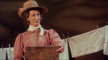 Movie still from “Oklahoma!” (1999), directed by Trevor Nunn – An older woman wearing a hat standing in front of a wooden box; Medium shot, Low angle