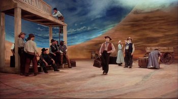 Movie still from “Oklahoma!” (1999), directed by Trevor Nunn – A group of people sitting and standing on a stage; Wide shot, Low angle