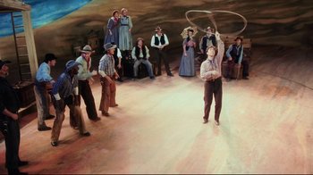 Movie still from “Oklahoma!” (1999), directed by Trevor Nunn – A group of people standing around a painting of a man holding a lasso; Wide shot, High angle