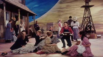 Movie still from “Oklahoma!” (1999), directed by Trevor Nunn – A group of people sitting on the ground in front of a painting; Wide shot, High angle