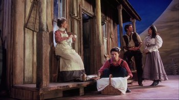 Movie still from “Oklahoma!” (1999), directed by Trevor Nunn – A woman sitting on a porch next to two men; Wide shot, Low angle