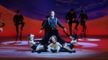 Movie still from “Oklahoma!” (1999), directed by Trevor Nunn – A group of people sitting on the ground with a man standing in front of them; Wide shot, Low angle