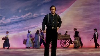 Movie still from “Oklahoma!” (1999), directed by Trevor Nunn – A man standing in front of a crowd of people on a stage; Wide shot, Low angle