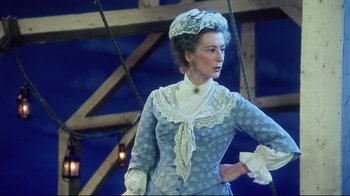 Movie still from “Oklahoma!” (1999), directed by Trevor Nunn – A woman dressed in period clothing standing in front of a wooden structure; Medium shot, Low angle