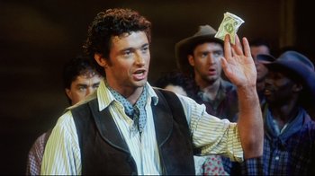 Movie still from “Oklahoma!” (1999), directed by Trevor Nunn – A group of men standing next to each other holding money; Medium shot, Over the shoulder angle