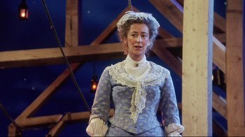 Movie still from “Oklahoma!” (1999), directed by Trevor Nunn – A woman dressed in old fashioned clothing standing in front of a wooden structure; Medium shot, Low angle