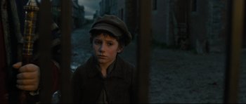 Movie still from “Oliver Twist” (2005), directed by Roman Polanski – A young boy wearing a hat standing on a street; Close Up shot, Over the shoulder angle