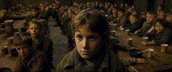 Movie still from “Oliver Twist” (2005), directed by Roman Polanski – A young boy is sitting in front of a group of people; Close Up shot, Over the shoulder angle