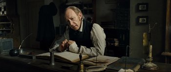 Movie still from “Oliver Twist” (2005), directed by Roman Polanski – An older man sitting at a desk with a book; Medium shot, High angle
