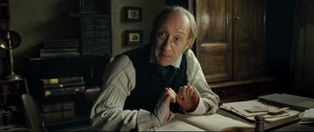 Movie still from “Oliver Twist” (2005), directed by Roman Polanski – An older man sitting at a table in front of a book; Medium shot, Low angle
