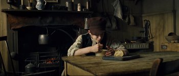 Movie still from “Oliver Twist” (2005), directed by Roman Polanski – A man sitting at a table with a loaf of bread in front of him; Medium shot, High angle