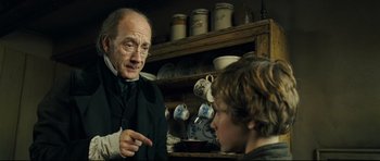 Movie still from “Oliver Twist” (2005), directed by Roman Polanski – A man and a boy are talking in front of a shelf; Medium shot, Over the shoulder angle