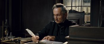 Movie still from “Oliver Twist” (2005), directed by Roman Polanski – An older man writing on a book in front of a window; Close Up shot, High angle