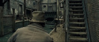 Movie still from “Oliver Twist” (2005), directed by Roman Polanski – A man wearing a hat standing next to a river; Wide shot, High angle