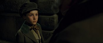 Movie still from “Oliver Twist” (2005), directed by Roman Polanski – A young boy wearing a suit and hat; Close Up shot, Over the shoulder angle