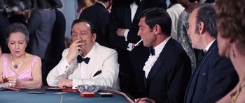 Movie still from “On Her Majesty's Secret Service” (1969), directed by Peter R. Hunt – Two men sitting at a table in formal wear smoking; Medium shot, Over the shoulder angle