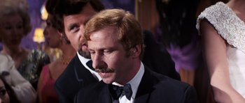 Movie still from “On Her Majesty's Secret Service” (1969), directed by Peter R. Hunt – A man in a suit and a bow tie looking down; Close Up shot, Over the shoulder angle