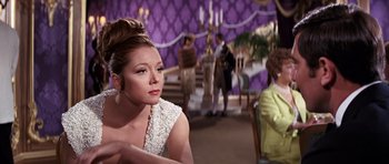 Movie still from “On Her Majesty's Secret Service” (1969), directed by Peter R. Hunt – A woman sitting in front of other people in a room; Medium shot, Over the shoulder angle