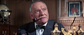 Movie still from “On Her Majesty's Secret Service” (1969), directed by Peter R. Hunt – An older man wearing a suit and tie with a pipe in his mouth; Close Up shot, Low angle