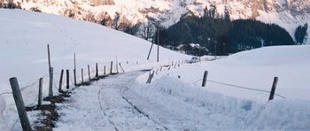 Movie still from “On Her Majesty's Secret Service” (1969), directed by Peter R. Hunt – A snowy road going down a hill with a car parked on the side of the road; Extreme Wide shot, High angle