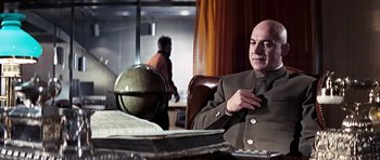 Movie still from “On Her Majesty's Secret Service” (1969), directed by Peter R. Hunt – A bald man in a brown suit sitting at a table; Medium shot, Low angle