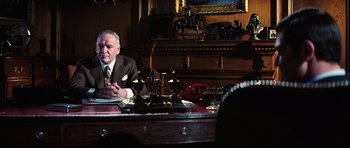Movie still from “On Her Majesty's Secret Service” (1969), directed by Peter R. Hunt – An older man sitting at a desk in front of a laptop computer; Medium shot, Low angle