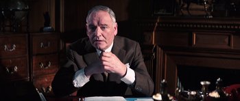 Movie still from “On Her Majesty's Secret Service” (1969), directed by Peter R. Hunt – An older man sitting at a table in front of papers; Close Up shot, Low angle