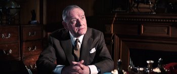 Movie still from “On Her Majesty's Secret Service” (1969), directed by Peter R. Hunt – An older man wearing a suit and tie sitting at a table; Medium shot, Low angle