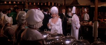 Movie still from “On a Clear Day You Can See Forever” (1970), directed by Vincente Minnelli – A woman in a white dress standing in front of a buffet line; Medium shot, Over the shoulder angle
