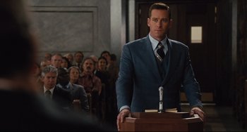 Movie still from “On the Basis of Sex” (2018), directed by Mimi Leder – A man in a blue suit stands at a podium in front of a crowd of onlookers; Medium shot, Low angle