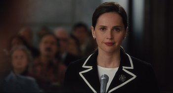 Movie still from “On the Basis of Sex” (2018), directed by Mimi Leder – A woman in a black and white jacket stands in front of a microphone; Close Up shot, Over the shoulder angle