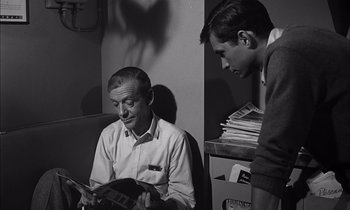 Movie still from “On the Beach” (1959), directed by Stanley Kramer – A man sitting next to another man holding a magazine; Medium shot, High angle